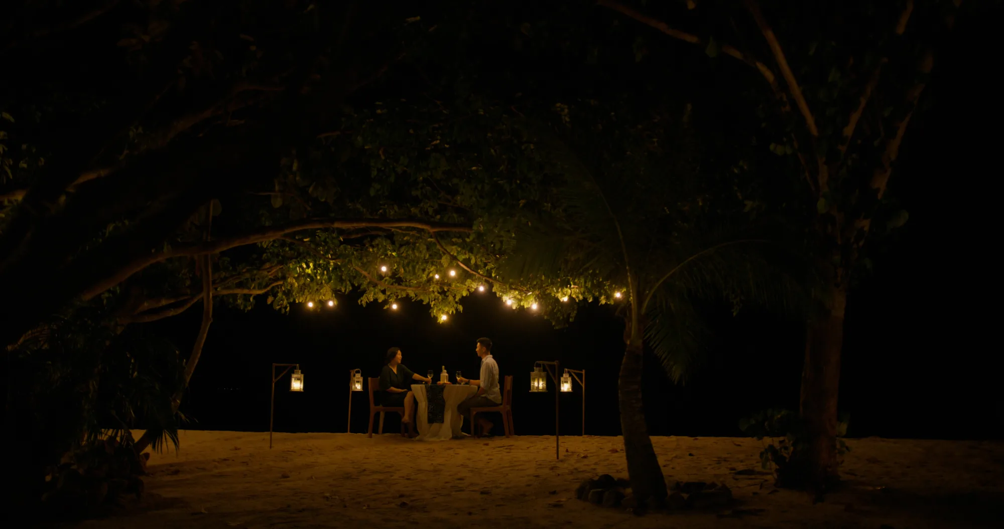 A private dinner table for two on the beach beneath lanterns and trees.
