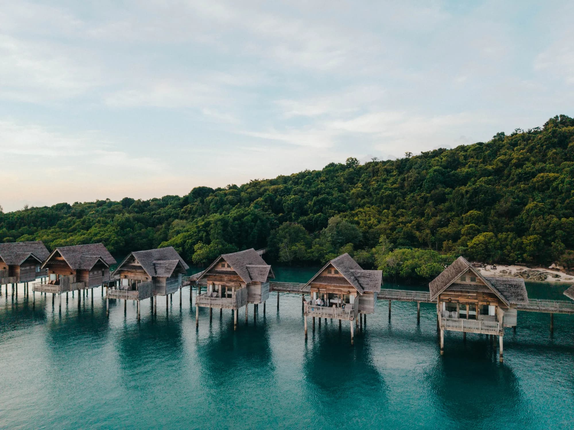 Aerial view of Telunas Private Island overwater villas beside forest and clear water.